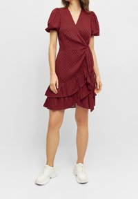 Robe portefeuille rouge à pois avec de courtes manches bouffantes, un ourlet volanté et un lien à la taille ; associée à des baskets blanches. Texture de tissu lisse.