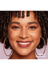 Prodotto labbra nella tonalità 07, un mauve caldo, su una persona con capelli ricci. Orecchini a cerchio dorati e un trucco occhi sottile completano il look.