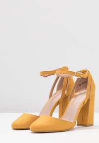 Żółte zamszowe szpilki z ostrym noskiem, paskiem w okolicy kostki i obcasem w kształcie bloku, z gładką wewnętrzną podszewką i minimalistycznym designem.