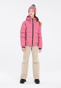 Giacca a vento rosa con cappuccio e dettagli neri, abbinata a pantaloni beige e stivali neri. Il modello indossa un berretto rosa abbinato con pompon.
