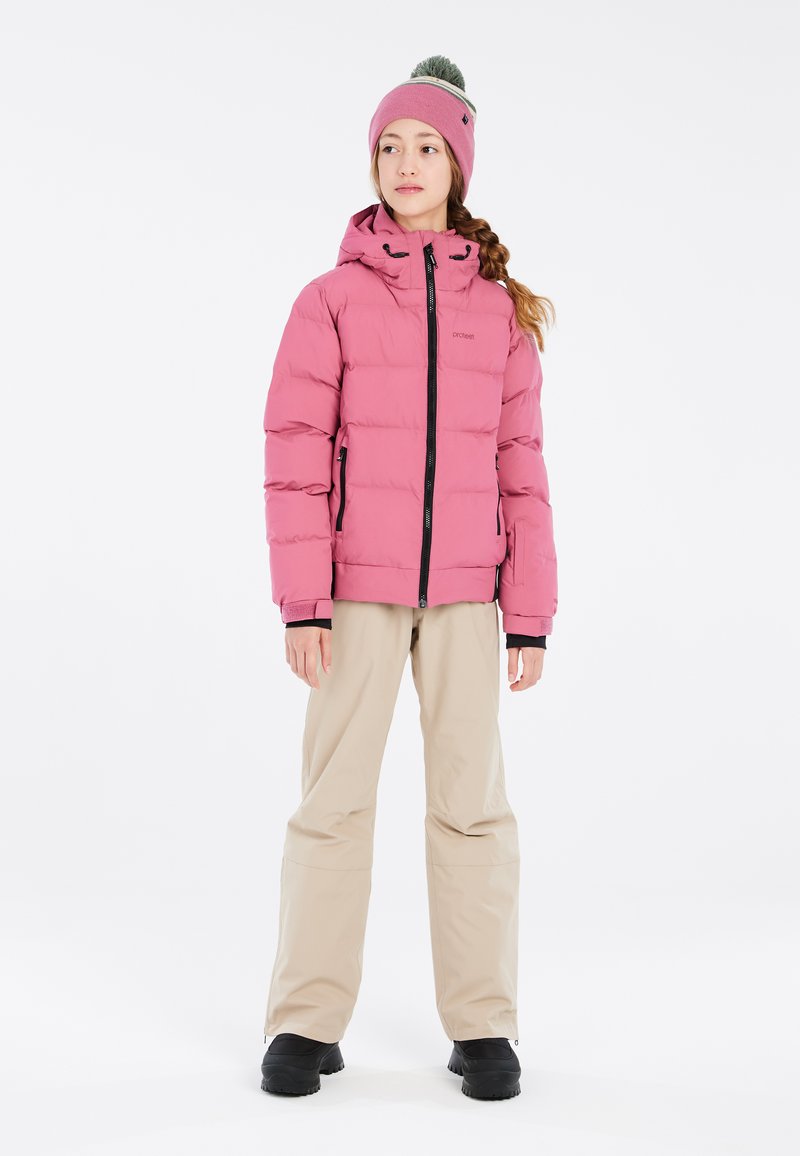 Giacca a vento rosa con cappuccio e dettagli neri, abbinata a pantaloni beige e stivali neri. Il modello indossa un berretto rosa abbinato con pompon.