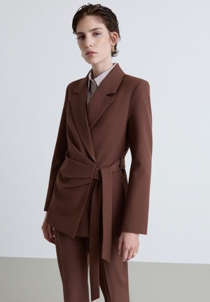 Person mit kurzen dunklen Haaren, die einen braunen Blazer mit Gürtel und passende Hosen vor einem einfarbigen hellen Hintergrund trägt.