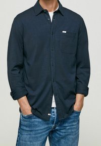 Camicia azzurra navy con bottoni, tasca, dettagli con etichetta bianca e maniche arrotolate, abbinata a jeans in denim azzurro chiaro.