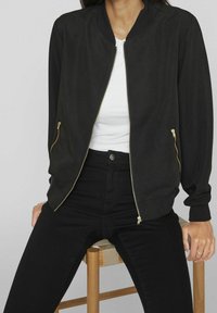 Zwarte bomberjack met gouden ritssluiting, lange mouwen en zijzakken, gecombineerd met een witte top en zwarte broek. Soepele textuur.