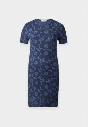 Femme portant une robe bleu marine à manches courtes, longueur genou, avec de grands motifs floraux foncés et des chaussures plates noires, debout devant un fond uni.