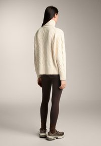Tömör krémszínű, kabelkötéses, garbós pulóver bordás mandzsettákkal, sötét leggings-szel, valamint szürke kiegészítőkkel ellátott barna sportcipővel párosítva.
