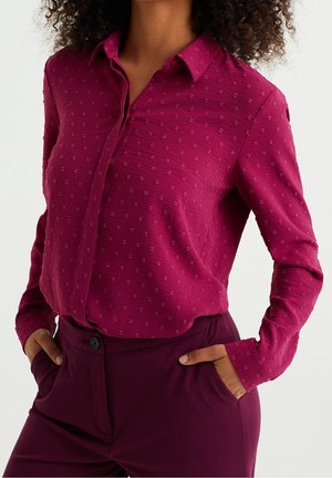 Fuchsia Hemd mit Knöpfen und feinem Strukturmuster, langen Ärmeln, Kragen und Manschetten. Kombiniert mit hochgeschnittenen Pflaumen-Hosen.