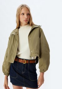 Olivengrøn cropped jakke med hætte, lynlåslukning og trykknapper. Bæret over en lys sweater og en denim mini-nederdel med et brunt bælte.