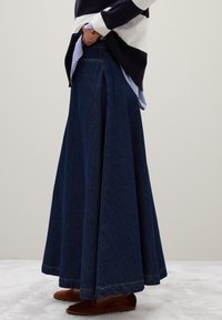 Personne portant une longue jupe en denim bleu foncé avec des chaussures plates marron, une chemise rayée et un pull noir et blanc, se tenant de profil.