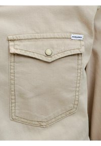Tasca sul petto in tessuto beige con patta e bottone, brandizzata con una piccola etichetta bianca con la scritta "Jack & Jones" sul bordo superiore destro.