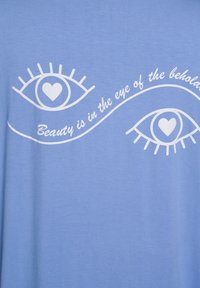 Vaaleansininen t-paita, jossa valkoinen graafinen kuva kahdesta tyylitellystä silmästä ja teksti "Kauneus on katsojan silmässä." Pehmeä koostumus.