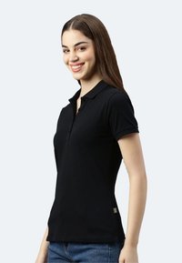 Junge Frau lächelnd, trägt ein eng anliegendes schwarzes Polo-Shirt mit kurzen Ärmeln und blaue Jeans, steht vor einem schlichten hellen Hintergrund.