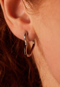 Zilveren ringoorbel in een gladde, gebogen vorm, dicht bij het oor. Het oppervlak is gepolijst met een reflecterende afwerking en minimale hardwaredetails.