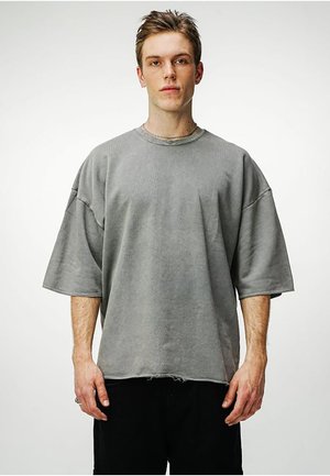Jonge man staat, draagt een oversized grijs T-shirt met korte mouwen en een versleten zoom en zwarte broek, neutrale uitdrukking, effen witte achtergrond.