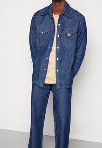 Jeansjacka med knappframsida, spetsig krage och två bröstfickor; mörkblå färg; matchad med en ljusbeige t-shirt och matchande byxor.