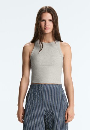 Jonge vrouw met lang bruin haar, gekleed in een grijs mouwloos cropped topje en blauwe gestreepte broek met hoge taille, staand tegen een lichte achtergrond.