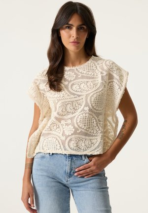 Woman with long brown hair wears a cream crochet short-sleeve top and light blue jeans, posing against a plain light background.