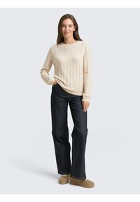 Helles beiges, geripptes Strickpullover mit langen Ärmeln, kombiniert mit dunklen Jeans und beigen Schuhen. Schlichtes Design mit Rundhalsausschnitt.