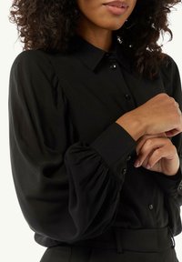 Black, long-sleeved blouse with a classic collar. Features puffed sleeves, buttoned cuffs, and a smooth, lightweight fabric texture.