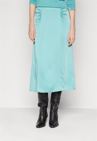 Une jupe midi turquoise avec des détails froncés sur les côtés, fabriquée dans un tissu lisse, assortie à des bottes noires ajustées jusqu'aux genoux.