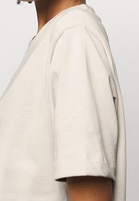 T-shirt in cotone beige con maniche corte, scollatura rotonda e dettagli di cucitura visibili. Il tessuto sembra liscio e leggero.