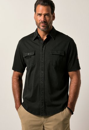 Man met baard die een zwart kort mouwen overhemd met borstzakken en beige broek draagt, staat met zijn handen in de zakken.