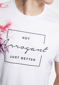 Camiseta blanca con un estampado rectangular negro que dice "NO Arrogante, SOLO MEJOR." Destacan patrones de humo en morado y rojo.