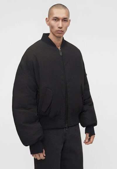 Άνδρας με κοντά μαλλιά που φοράει μαύρο υπερμεγέθες bomber μπουφάν με φερμουάρ και μαύρο παντελόνι, κοιτώντας ευθεία μπροστά με ουδέτερη έκφραση.