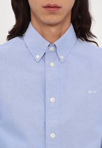 Homme portant une chemise bleu clair à boutons avec un col boutonné et le logo « NN.07 » brodé sur la poitrine.