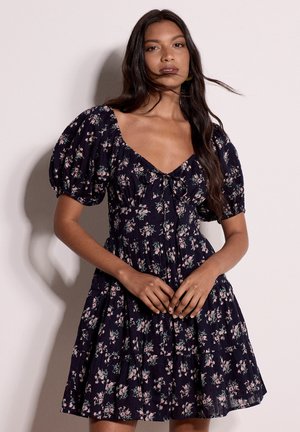 Woman stands against a plain background wearing a dark floral dress with puffed short sleeves and a V-neckline, hands clasped in front.