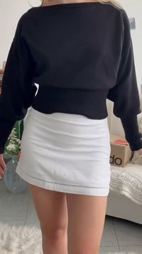 Άτομο που φοράει μαύρο μακρυμάνικο crop top και λευκή μίνι φούστα, στέκεται μέσα σε εσωτερικό χώρο κοντά σε έναν καναπέ και ένα κουτί.
