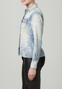 Camicia di denim azzurro chiaro con un motivo tie-dye sbiadito, abbottonatura frontale e due tasche sul petto; indossata con leggings neri.