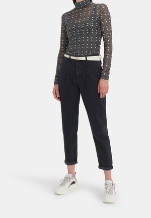 Person iført sort mønstret bluse med lange ærmer, sorte højtaljede opsmøgede jeans med hvidt bælte og hvide chunky sneakers, der står mod hvid baggrund.