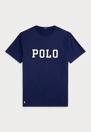 Jūras zilas kokvilnas T-krekls ar īsām piedurknēm, kuram ir liels balts "POLO" logotips drosmīgā fontā un mazs logotipa elements uz apakšmalas.