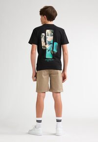 Camiseta negra con un gráfico en forma de palma que muestra una escena de playa y océano, combinada con pantalones cortos beige y zapatillas blancas.