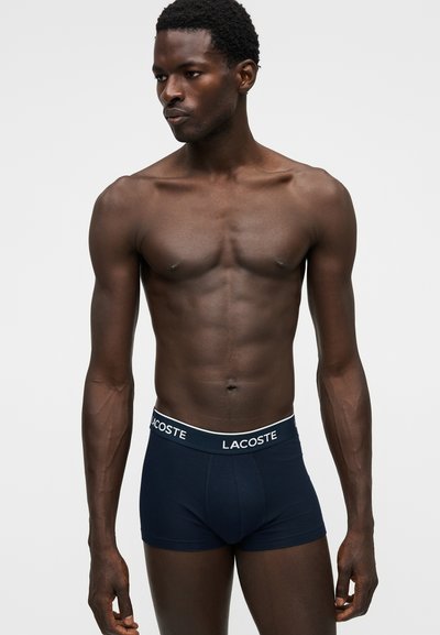 Мускулест тъмнокож мъж, носещ тъмносини боксерки Lacoste, стои на обикновен светъл фон, гледайки наляво.