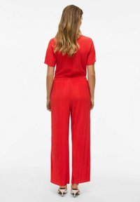 Mono rojo plisado con mangas cortas, cintura elástica, pantalones de pierna ancha y textura suave. Presenta una parte superior ajustada y fluye suavemente.