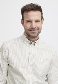 Lys grønn og hvitstripet skjorte med knapper og brystlomme; har subtile logoaksenter i rødt og blått. Glatt stofftekstur.