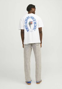 Camiseta de algodón blanca con un gráfico de un pájaro colorido en la parte posterior que dice "PORTO ALEGRE. SINCE MCMXC" en azul, combinada con pantalones anchos de mezclilla gris claro.