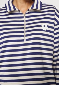 Randig tröja i marinblått och creme, med en halvdragkedja och en vit blommig logotyp på vänster bröst. Slät textur.