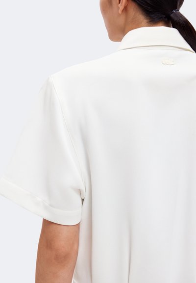 Femme aux cheveux foncés attachés, portant une chemise blanche à manches courtes avec col, vue de dos et de côté sur un fond clair.