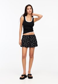 Schwarzes Tanktop, das über einem weißen Tanktop getragen wird, kombiniert mit einem schwarzen, gepunkteten, gestuften Minirock und schwarzen flachen Sandalen. Einfaches Design und lässiger Stil.