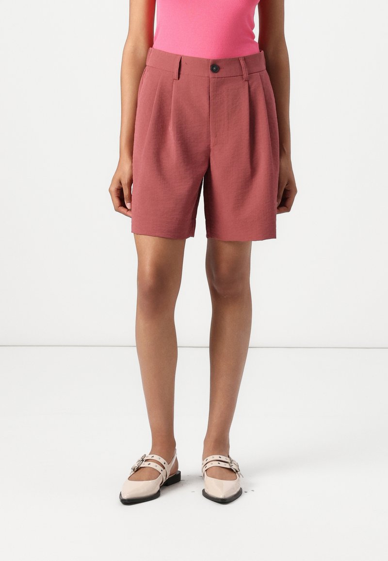 Shorts taillés rose avec une taille ceinturée, des plis avant, des poches latérales et une texture lisse ; associés à des chaussures plates nud.