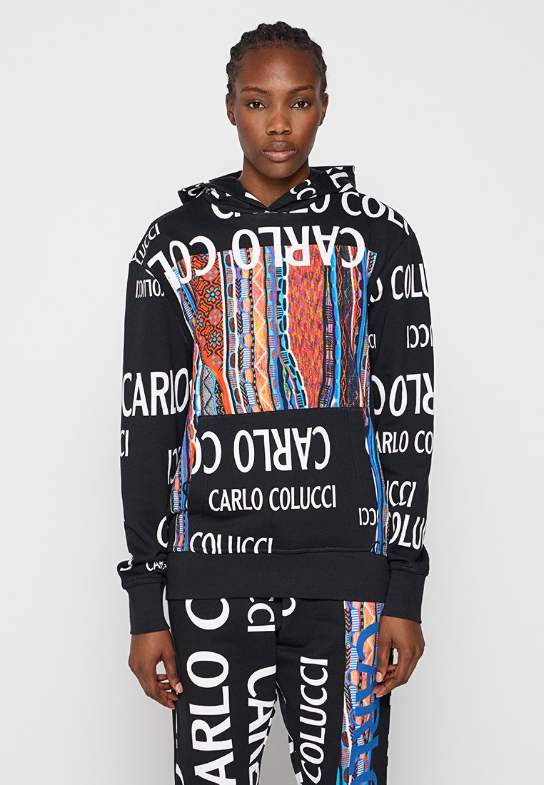 Joven vistiendo una sudadera negra con capucha y pantalones con estampados multicolores audaces y texto blanco "Carlo Colucci" impreso por todas partes.