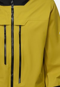 Giacca resistente all'acqua di colore giallo brillante con colletto nero. Presenta due tasche con zip e una finitura liscia e opaca.