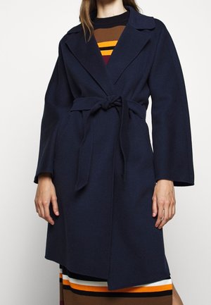 Frau trägt einen marineblauen Mantel mit Gürtel über einem gestreiften Rock in Braun, Orange, Gelb und Weiß vor einem schlichten hellen Hintergrund.