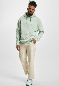 Felpa verde chiaro con tasca a marsupio, indossata su pantaloni cargo color crema. Il modello indossa sneaker bianche con accenti verdi.