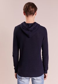 Námorně modrá pletená mikina s kapucí, která má texturovaný design, dlouhé rukávy a kontrastní kapuci. Upnutý pas a manžety.