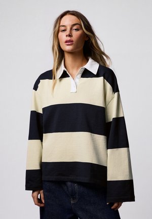 Femme portant un maillot de rugby à manches longues avec de larges rayures horizontales noires et beiges et un col blanc, debout devant un fond uni.