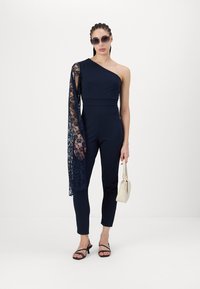 Mornarski kombinezon z enim ramenom, z čipkastim rokavom. Prilagojena zasnova, ožji hlačnic, v kombinaciji z črnimi sandali s trakovi in belim ročnim torbicami.